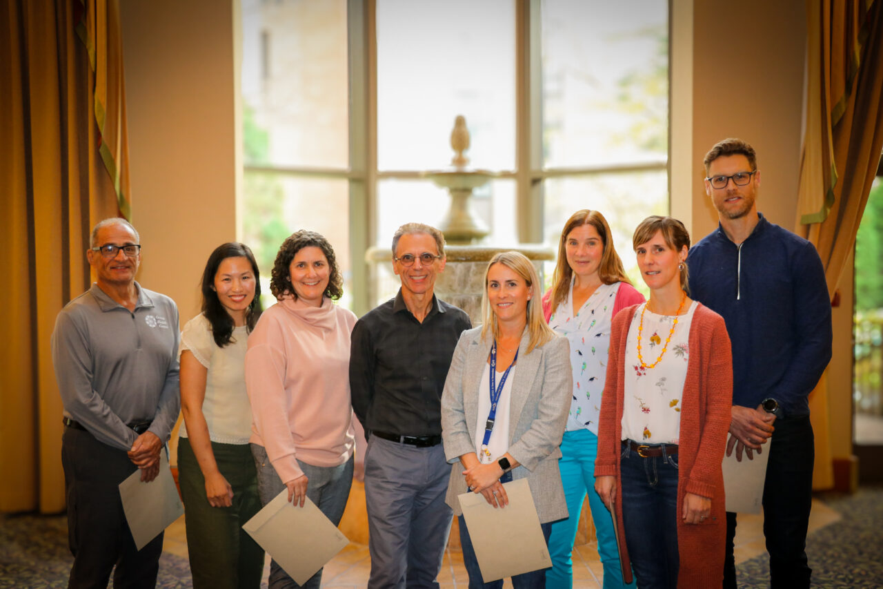 Group photo of eight GHC physicians, some holding certificates, standing indoors in a well-lit room with a decorative fountain in the background.